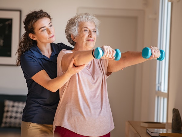 Older woman with therapist doing weighted exercise lifting arms in front of her body while holding dumbells (Source: Shutterstock)